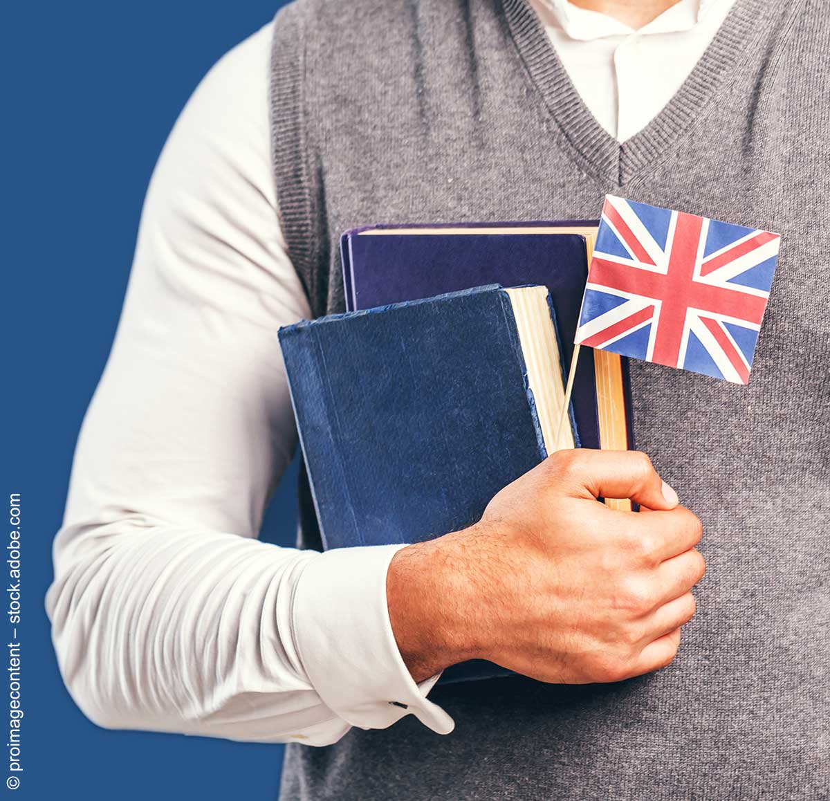 Ein Mann in weißem Hemd und grauem Pullunder trägt zwei Bücher und eine kleine Großbritannien-Flagge vorm Oberkörper