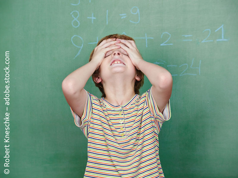 Eine Junge vor einer Schultafel, der sich wegen mathematischer Rechenaufgaben die Haare rauft.